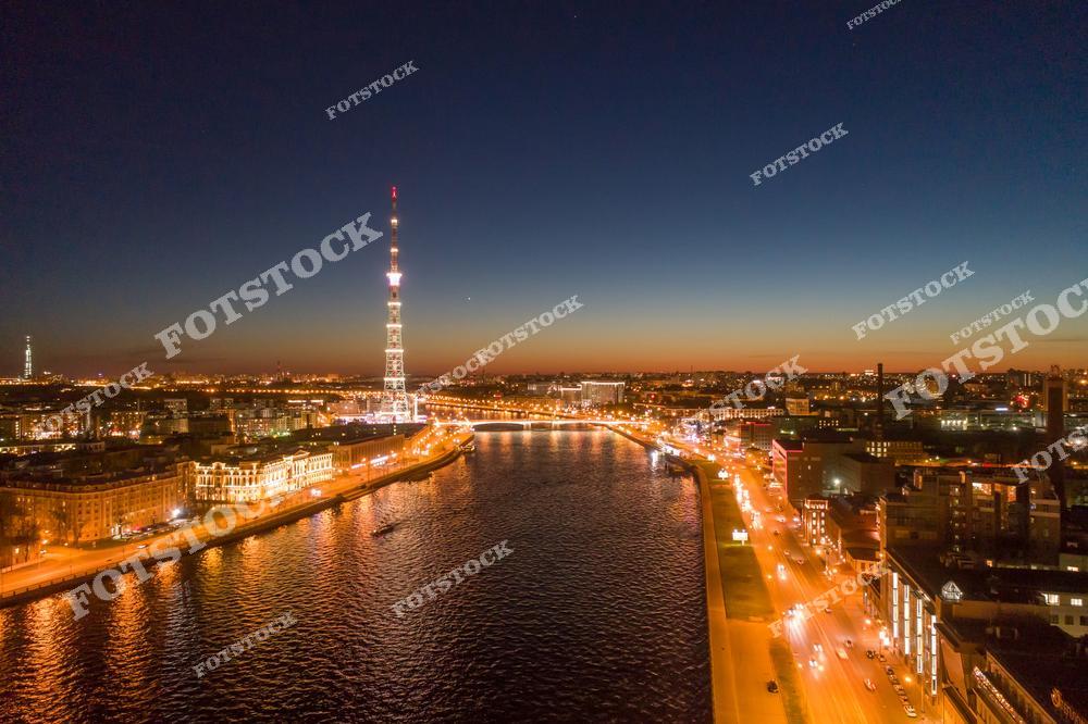Панорамная фотография Санкт-Петербурга ночью с высоты птичьего полета. На снимке видна река Нева, подсвеченные набережные, мосты и городские улицы с вечерними огнями. Съемка с дрона позволяет оценить масштабы городской инфраструктуры, архитектурные ансамбли и красоту ночного города. Фотография передает атмосферу ночного Петербурга, величие его городской среды и панорамные виды исторического центра. Снимок идеально подходит для туристических публикаций, фотостоков, рекламы, оформления интерьеров и материалов о достопримечательностях Северной столицы России.